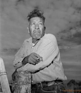 A man leans on a fencepost in a black-and-white photograph from the 1940s. He is wearing casual clothing and looks off into the distance, unsmiling.